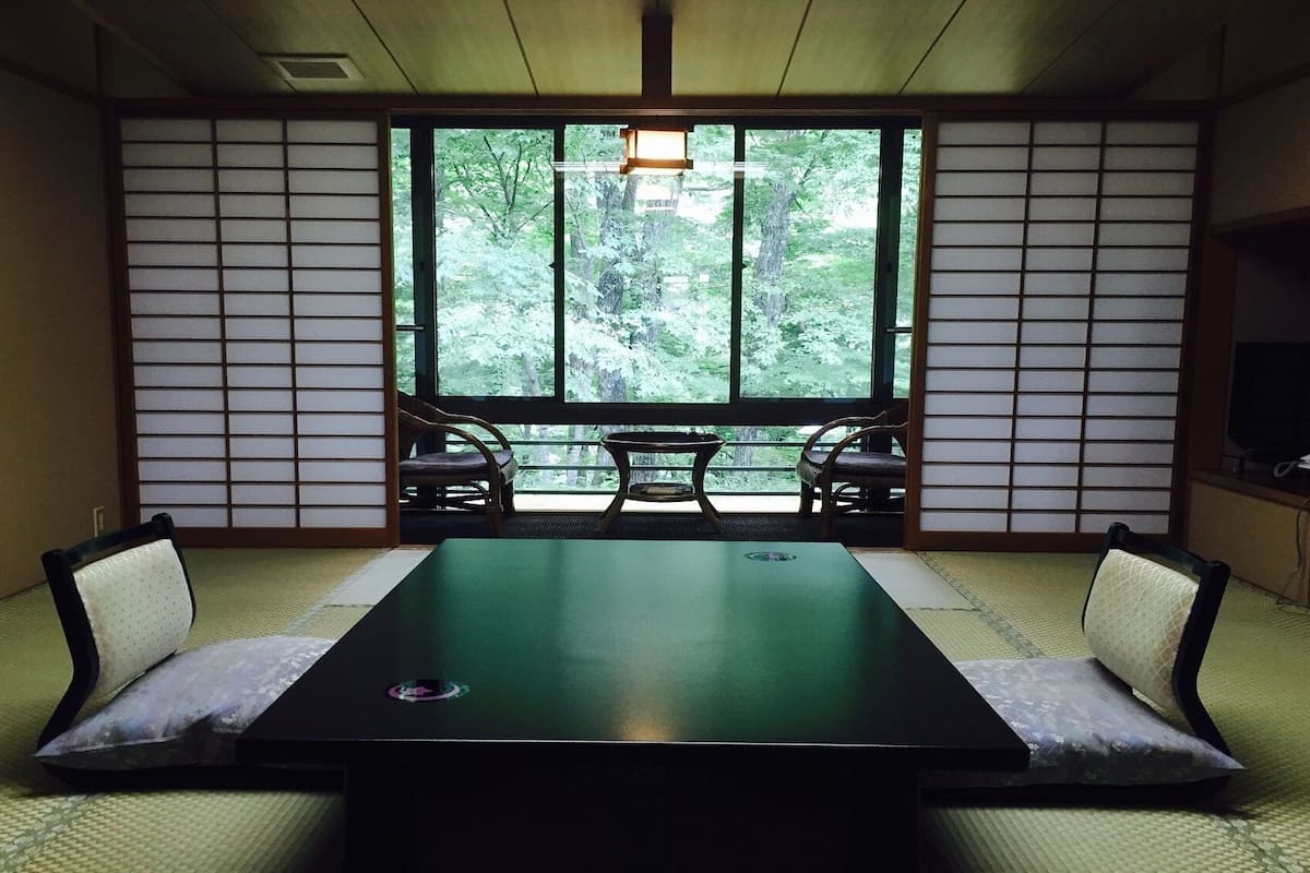 Ein traditioneller japanischer Raum mit Tatami-Bodenbelag, einem niedrigen Tisch und Blick auf einen Wald durch große Fenster.