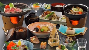 Diversos pratos e panelas japonesas, incluindo sashimi, legumes e tofu, dispostos sobre uma mesa com hashis e um copo de água.