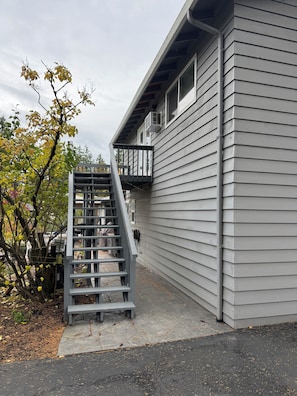 Un edificio residencial con fachada gris, una escalera de madera que conduce a un balcón, y un árbol con hojas amarillas al fondo.