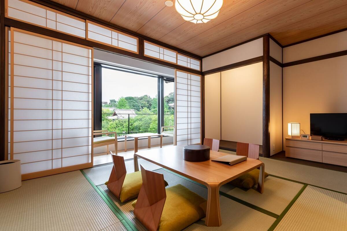 Ein traditioneller japanischer Raum mit Tatami-Bodenbelag, einem niedrigen Tisch, Kissen, einem Fernseher und Schiebetüren.