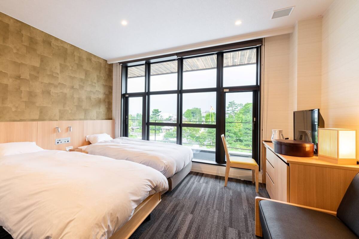 Ein Hotelzimmer mit zwei Betten, einem großen Fenster, einem Schreibtisch mit Lampe und einem Fernseher.