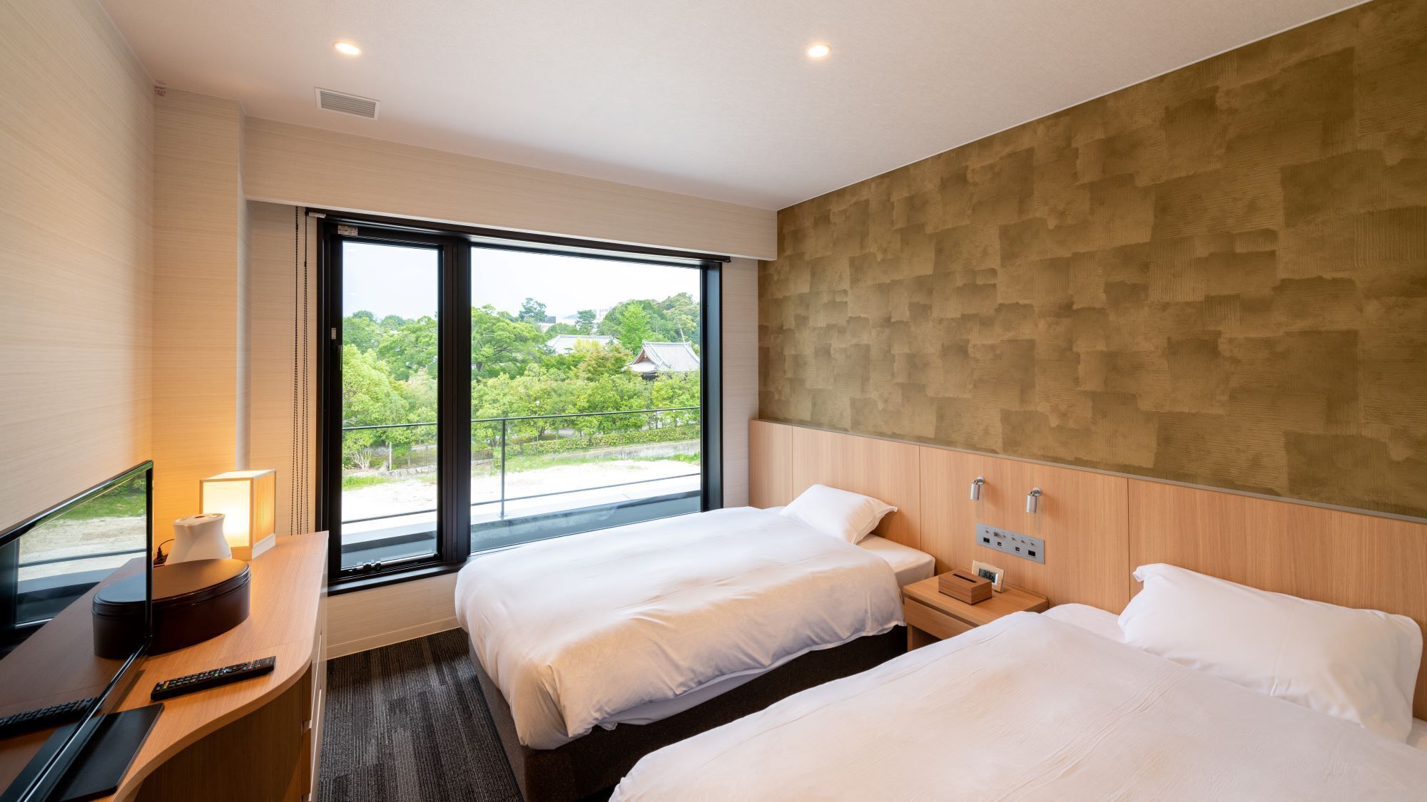 A hotel room with two beds, a desk with a TV, and a large window offering a view of greenery.