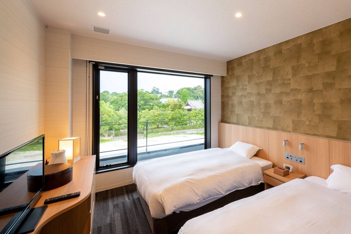 Ein Hotelzimmer mit zwei Betten, einem Schreibtisch mit Fernseher und einem großen Fenster mit Blick auf Grünflächen.
