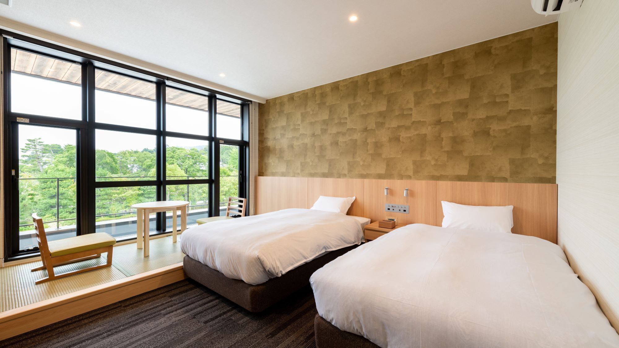 A hotel room with two beds, a small table, a chair, and a large window offering a view of greenery.
