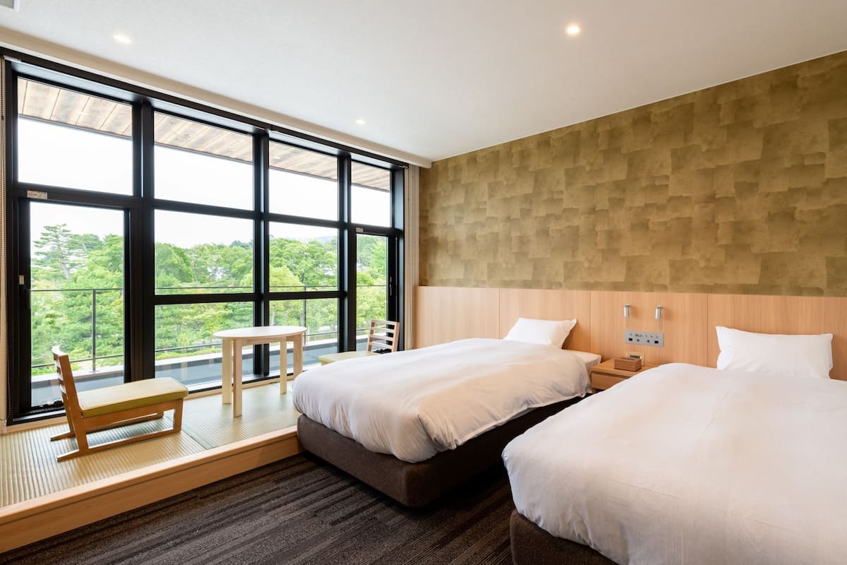 Ein Hotelzimmer mit zwei Betten, einem kleinen Tisch, einem Stuhl und einem großen Fenster mit Blick auf Grünflächen.