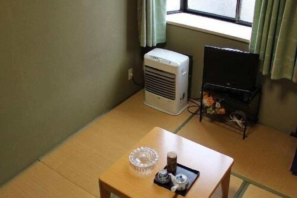 A room with tatami flooring, a small wooden table with a glass bowl and a teapot, a white air conditioner, and a television on a stand.