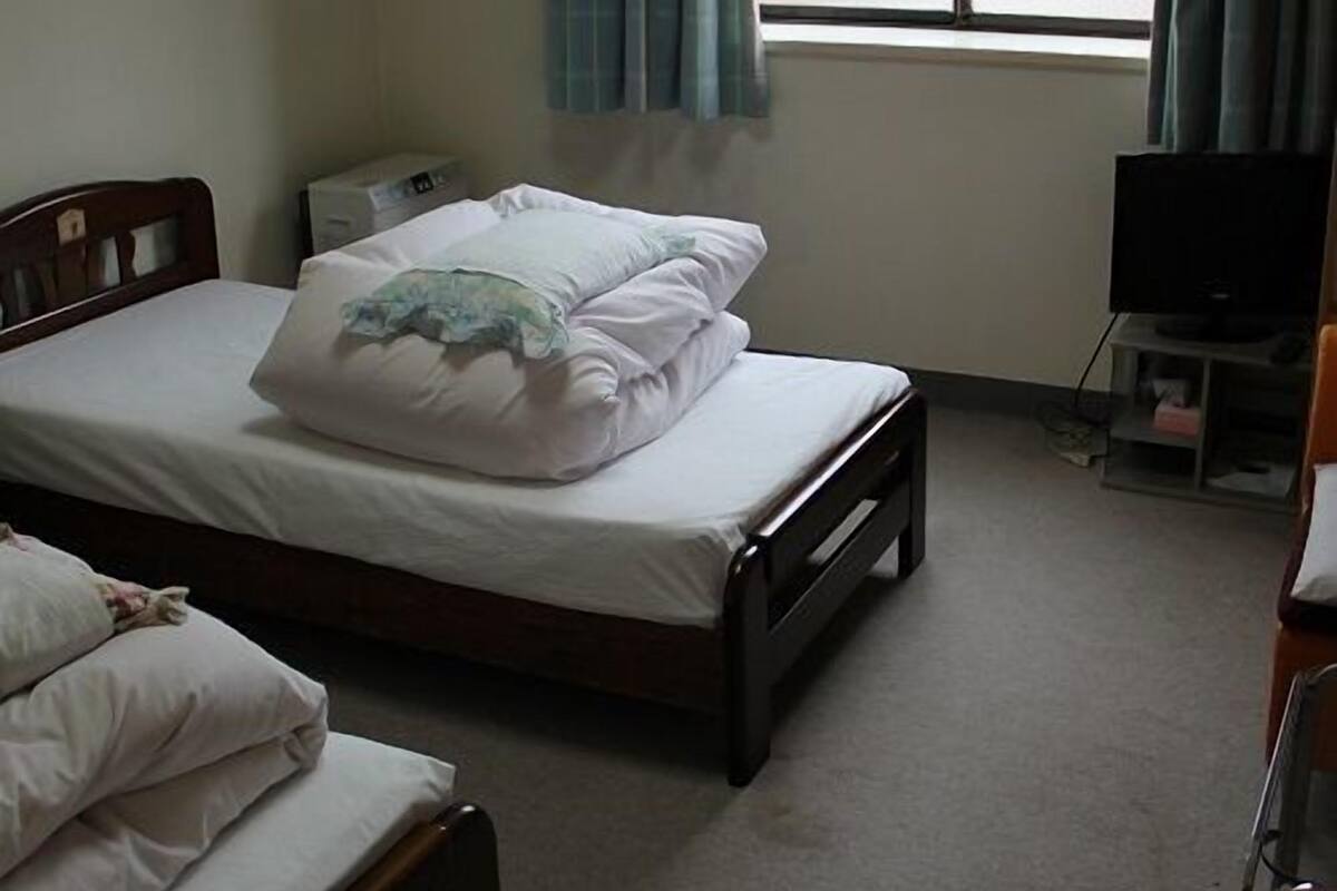 A single bed with a wooden frame, a bedside table, a television on a stand, and a window with curtains.