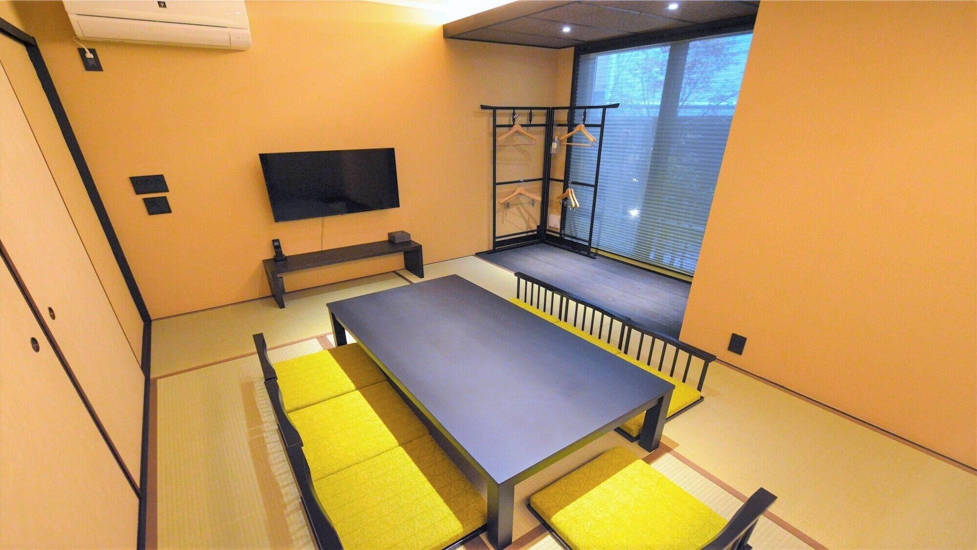 A room with a long table, benches, a TV, and a window with blinds.