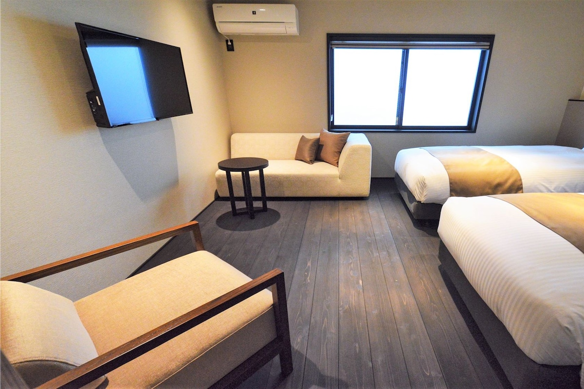 A hotel room with a sofa, two single beds, a small table, a TV, and a window.