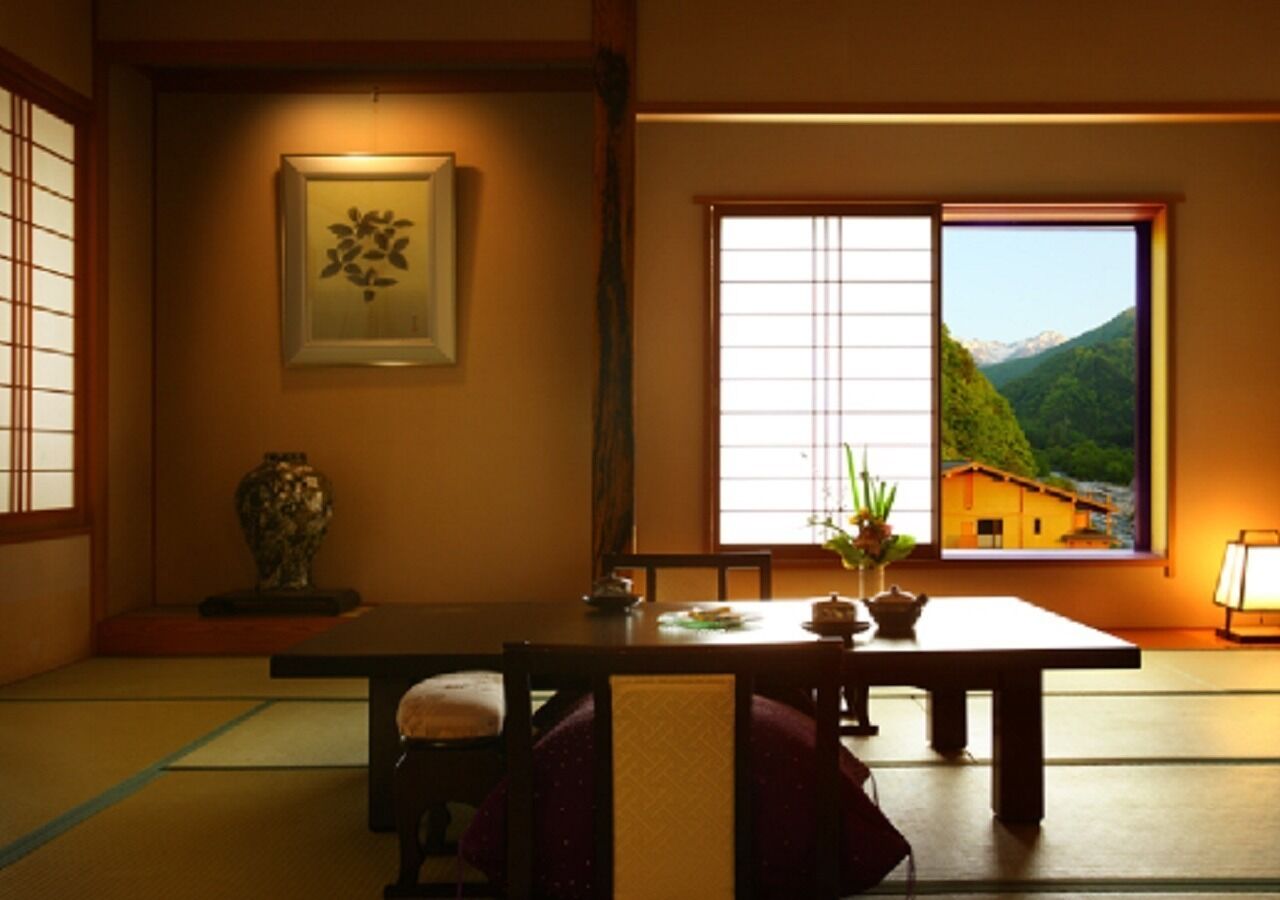 Una habitación tradicional japonesa con suelo de tatami, una mesa baja, una obra de arte enmarcada y un ventanal con vistas a la montaña.