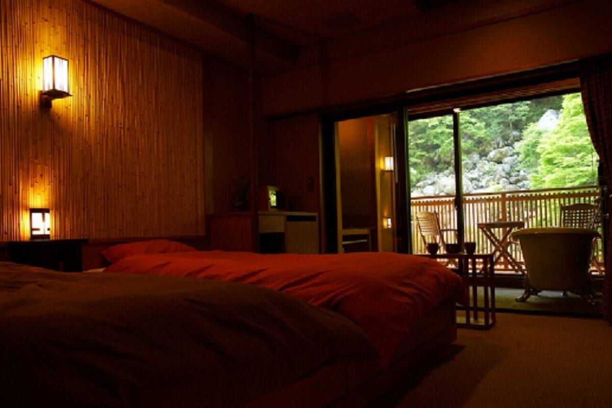 A bedroom with a large bed, a wooden wall, and a sliding door leading to an outdoor area with a chair and a bathtub.