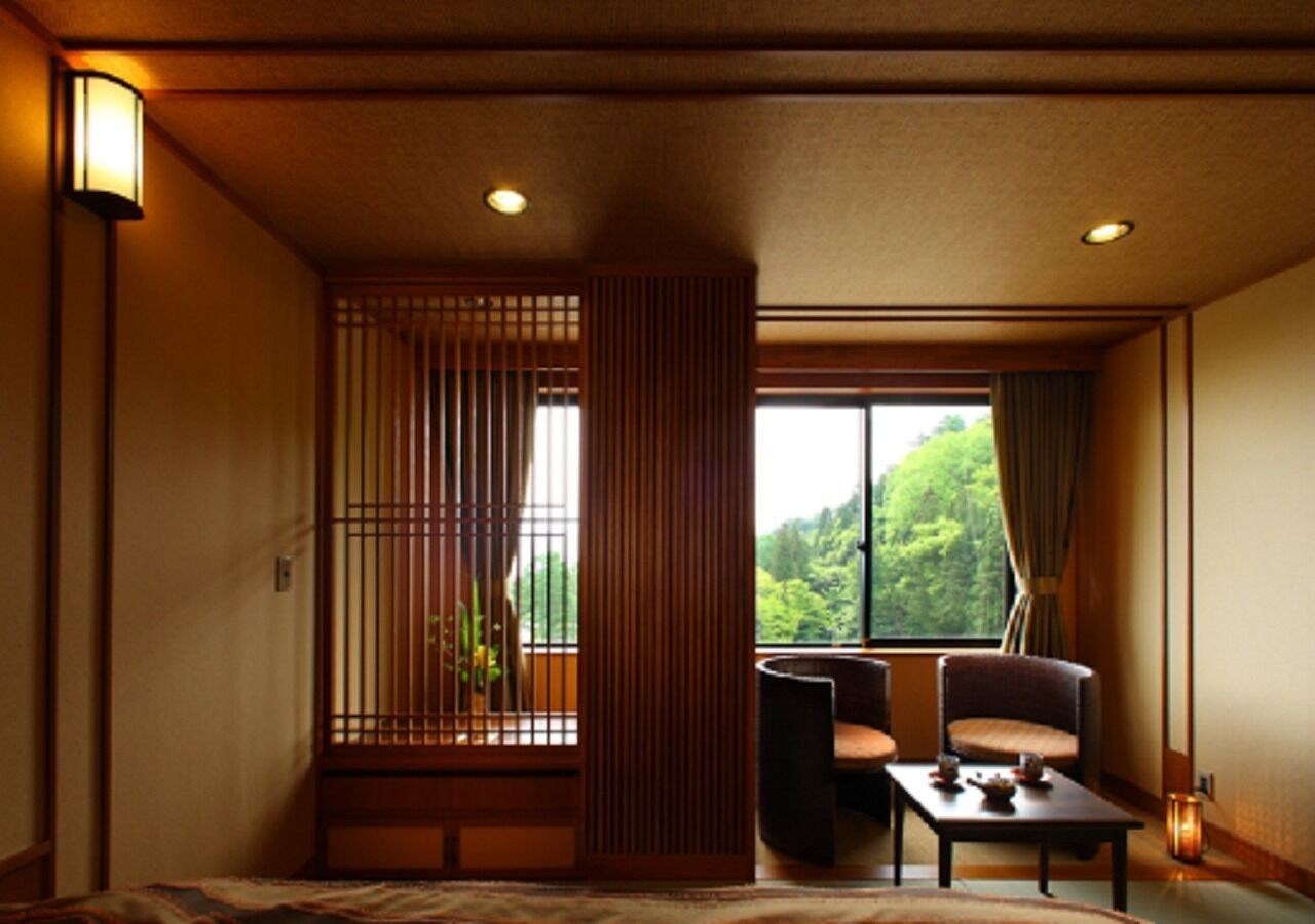Una habitación japonesa tradicional con suelo de tatami, una mesa baja con cojín, una silla y una ventana con vistas a la vegetación.