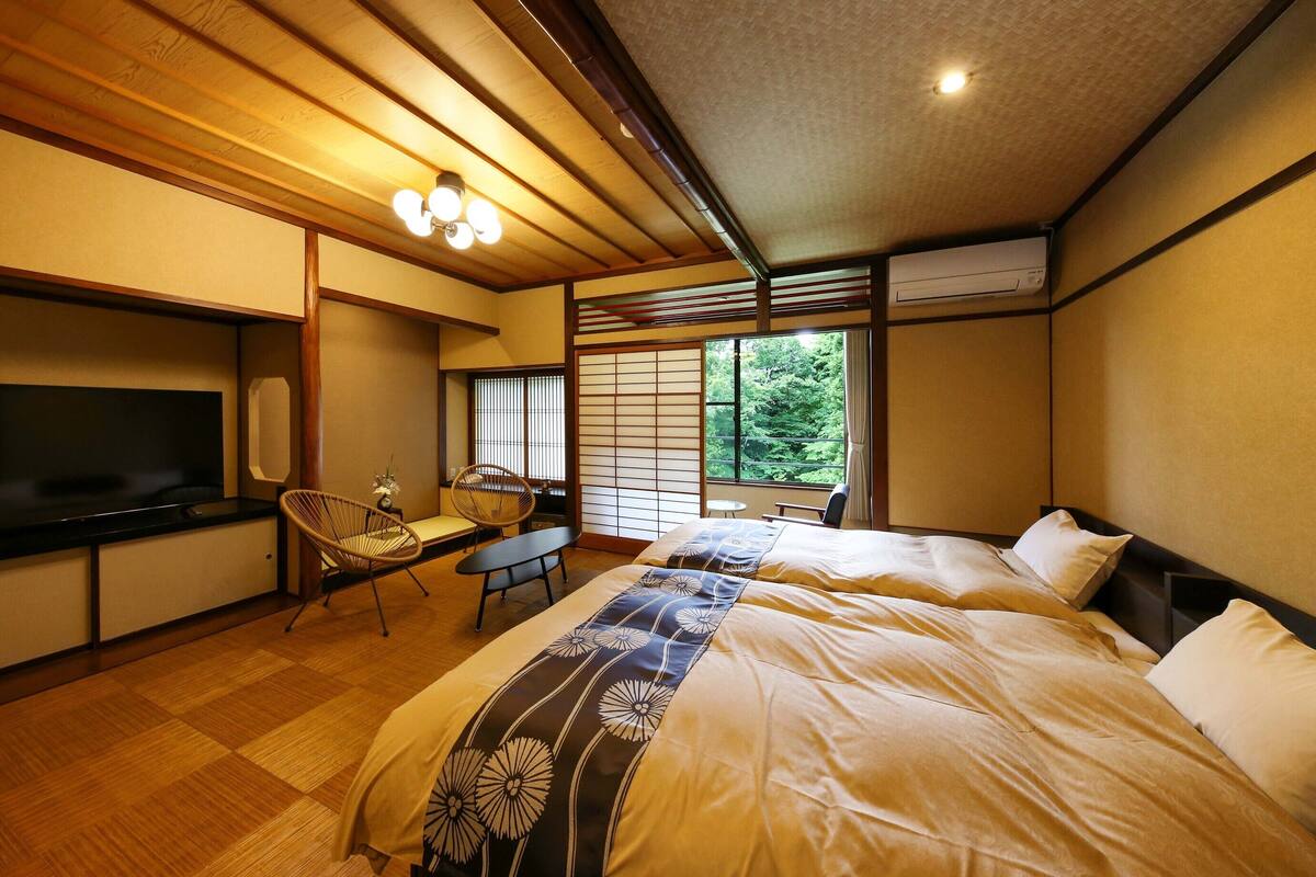 A traditional Japanese-style bedroom with tatami flooring, a television, a small table, and a view of greenery through the window.