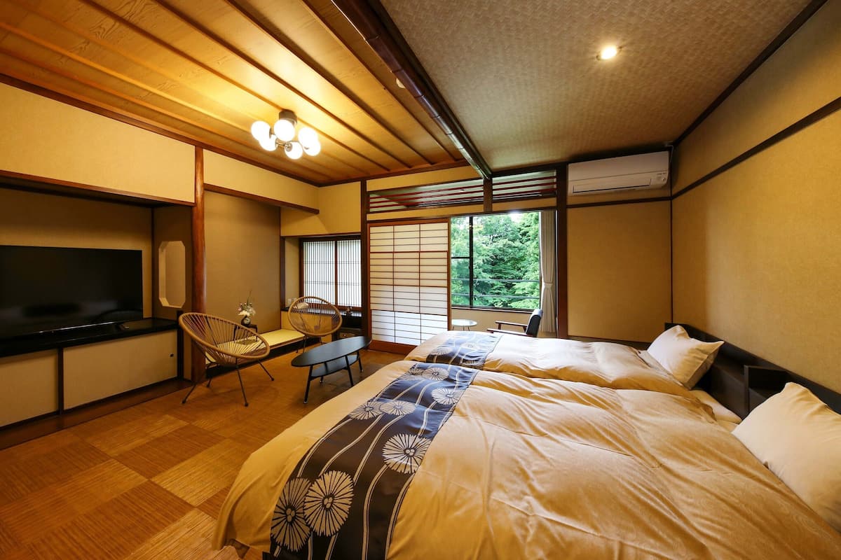 Un dormitorio de estilo japonés tradicional con suelo de tatami, un televisor, una mesita y vistas a zonas verdes por la ventana.