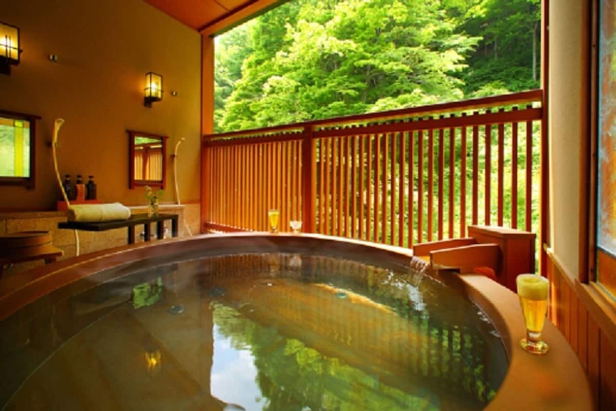 A hot tub with a wooden deck, a glass of beer, and a view of a forest.