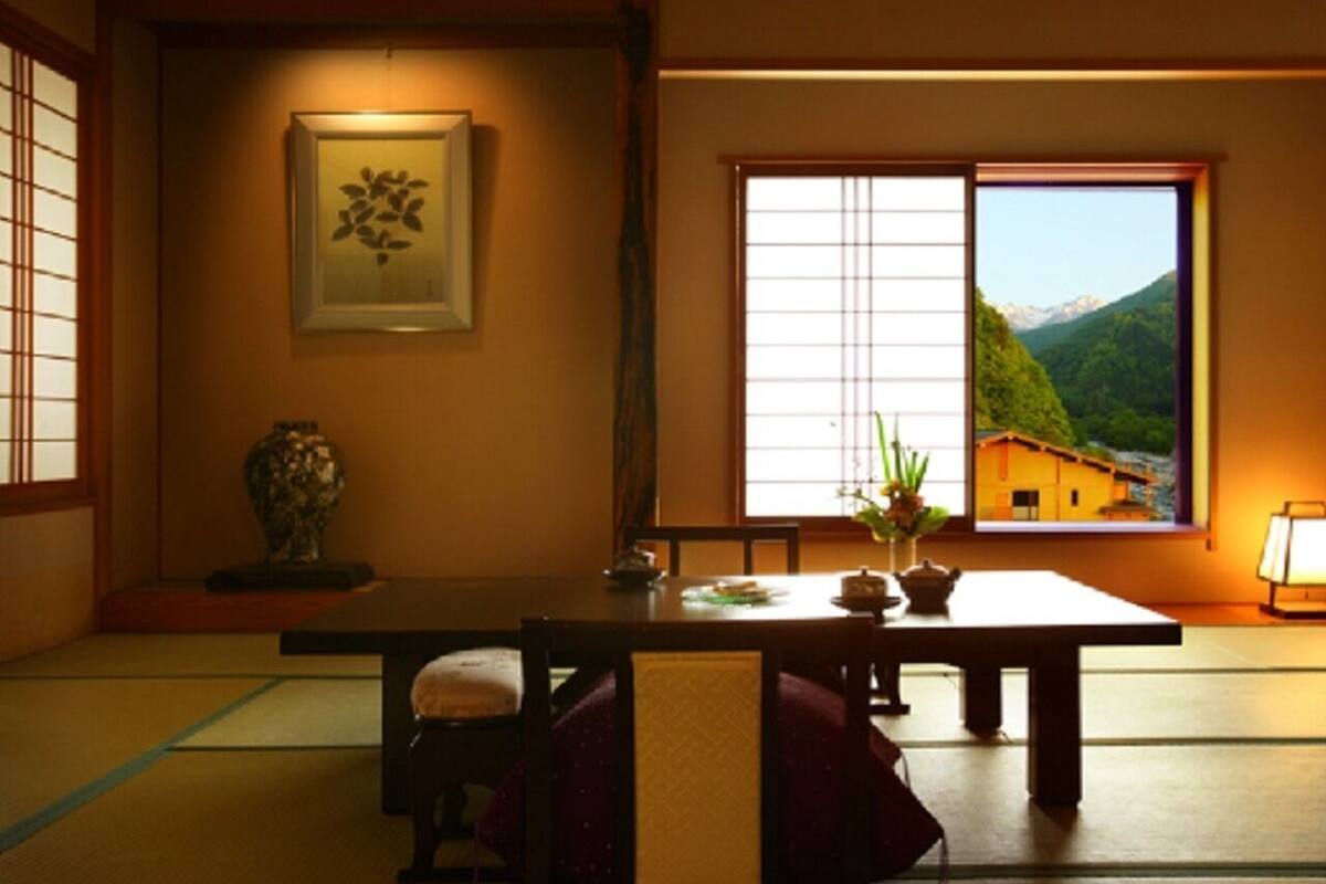 A traditional Japanese room with tatami flooring, a low table, a framed artwork, and a window with a mountain view.