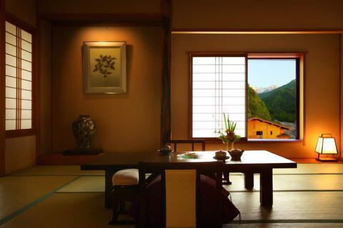 A traditional Japanese room with tatami flooring, a low table, a chair, a framed picture, a window with a view of a mountain, and a lamp.