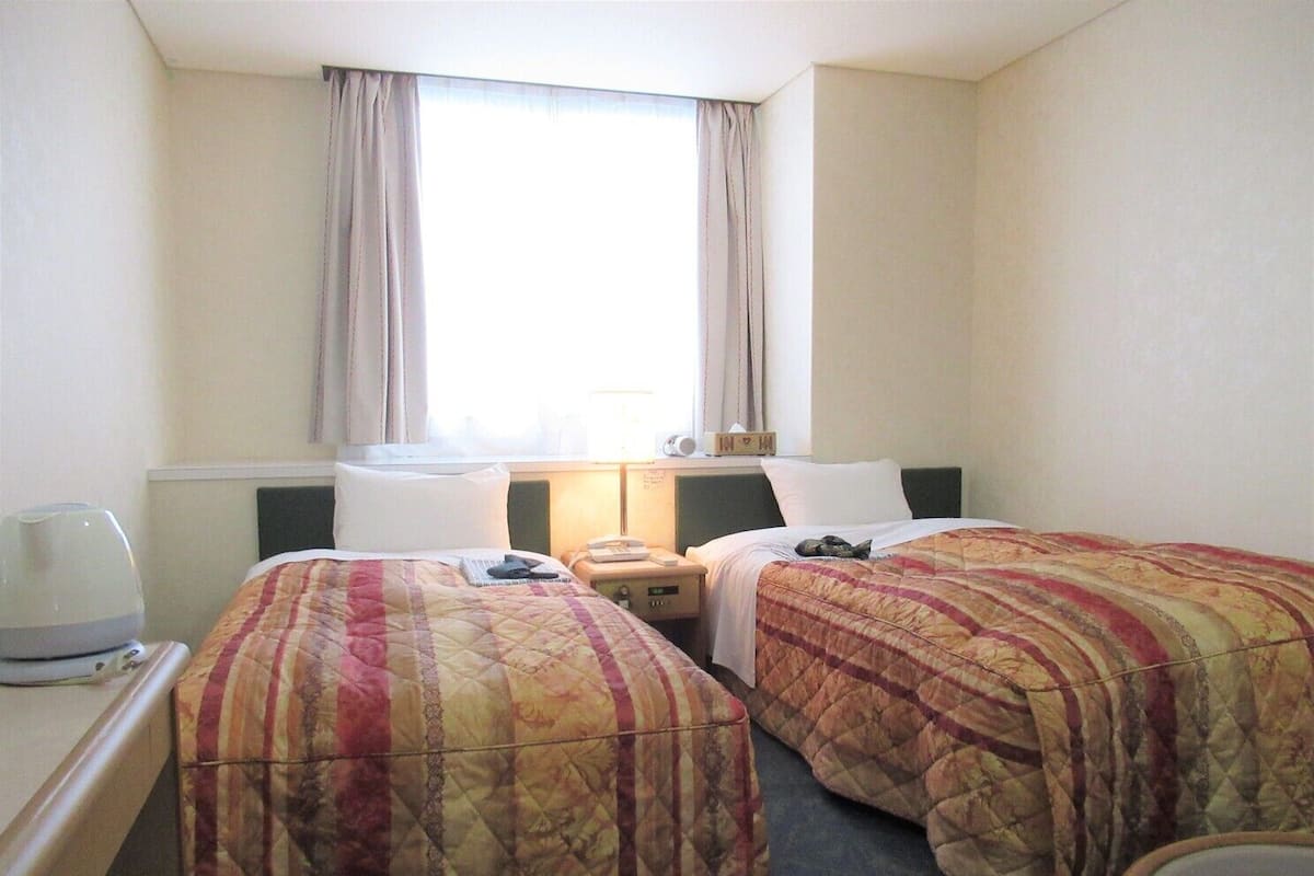 A hotel room with two beds, a desk with a lamp, a bedside table with a phone, and a window with curtains.