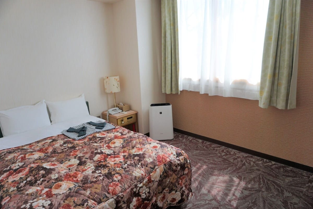 A hotel room with a bed, a bedside table with a lamp, a telephone, and a small refrigerator.
