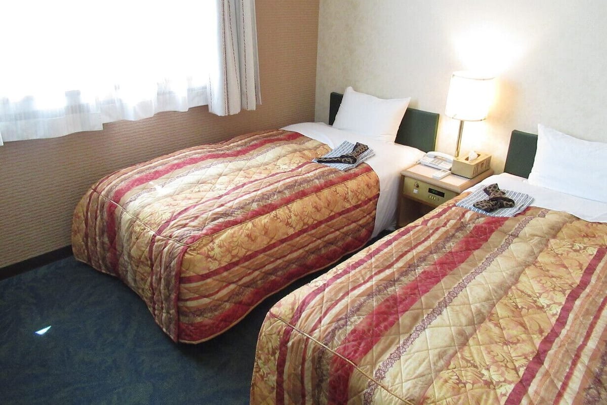 A hotel room with two beds, a nightstand, a lamp, and a window with curtains.