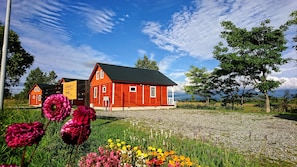 A red wooden house with a black roof, surrounded by a gravel driveway and a grassy area with flowers.