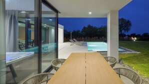 Un espacio moderno para comer al aire libre con piscina, paredes de vidrio y vistas al jardín.
