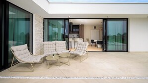 Un espacio exterior moderno amueblado con un conjunto de sofás y una mesita de centro, una cocina al fondo y un ventanal de vidrio que da a un balcón.