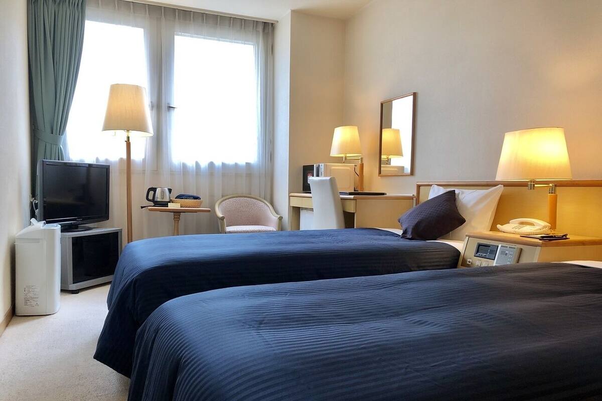 A hotel room with two beds, a TV, a desk with a lamp, a chair, and a bedside table with a phone.