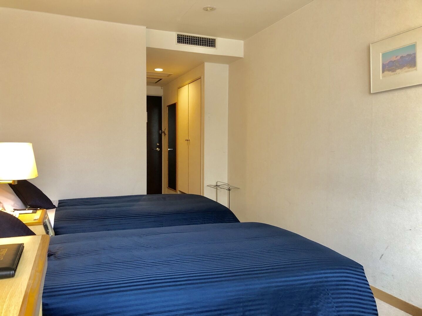 A hotel room with two beds, a desk with a lamp, and a framed picture on the wall.