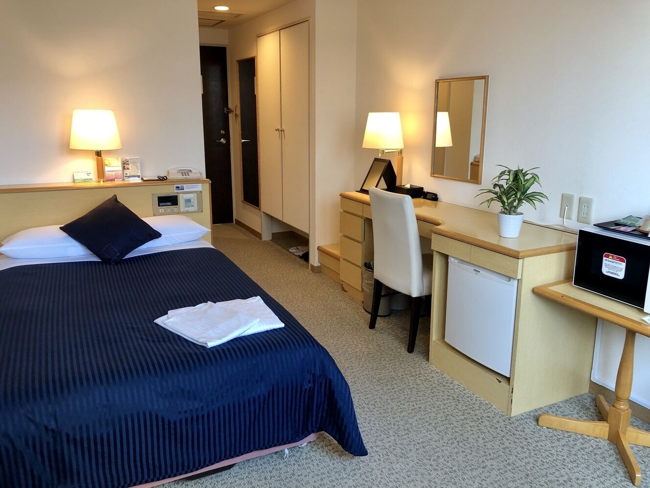 A hotel room with two beds, a desk, a chair, a small refrigerator, and a bedside table with a lamp.