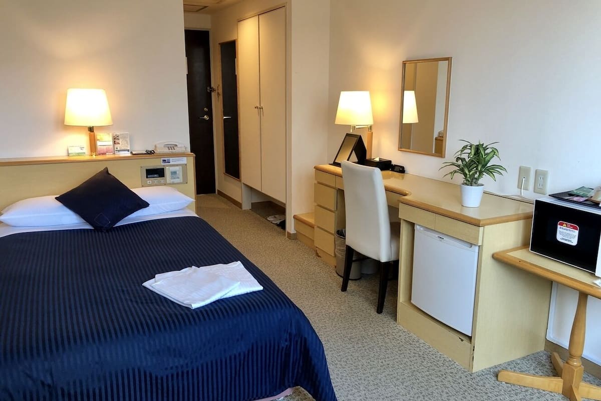 A hotel room with two beds, a desk, a chair, a small refrigerator, and a bedside table with a lamp.