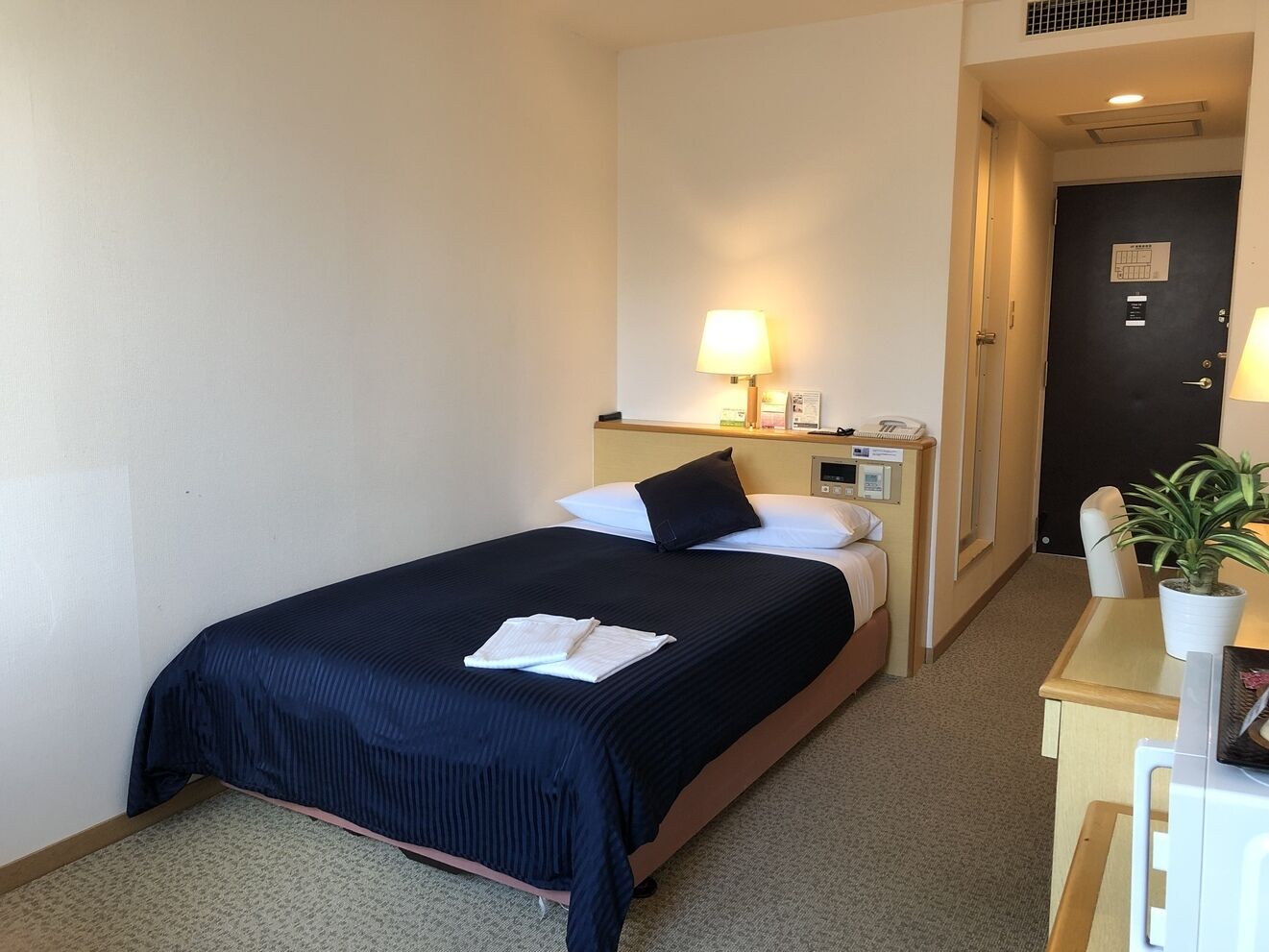 A hotel room with a bed, bedside table, lamp, and a small desk with a chair and a potted plant.