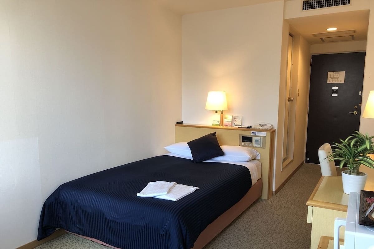 A hotel room with a bed, bedside table, lamp, and a small desk with a chair and a potted plant.