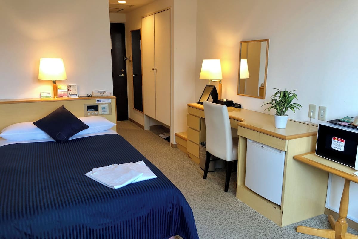 A hotel room with a bed, desk, chair, and a small refrigerator.