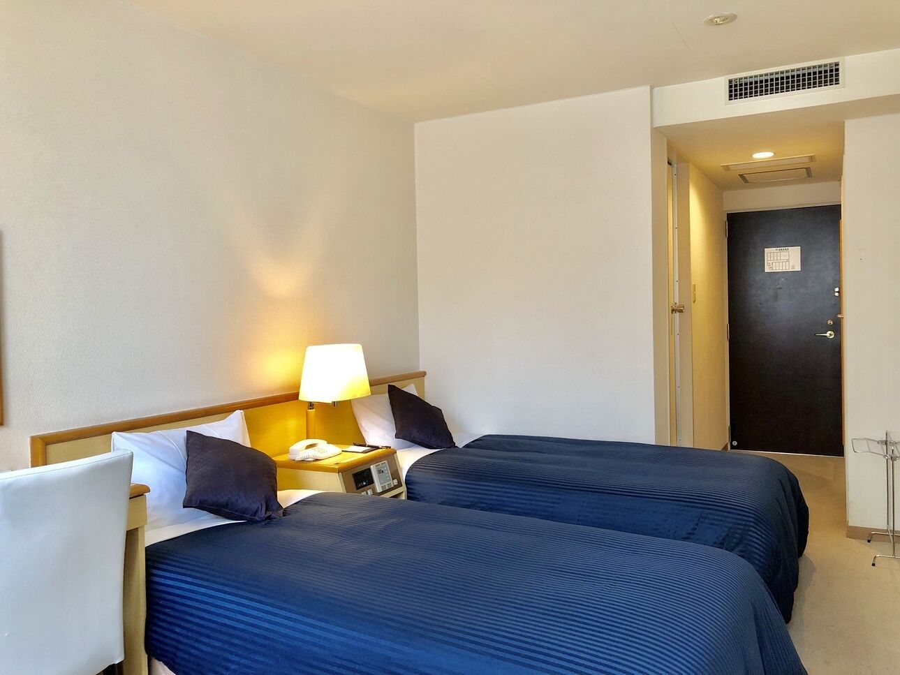 A hotel room with two beds, a nightstand with a lamp, a phone, and a door with a lock.
