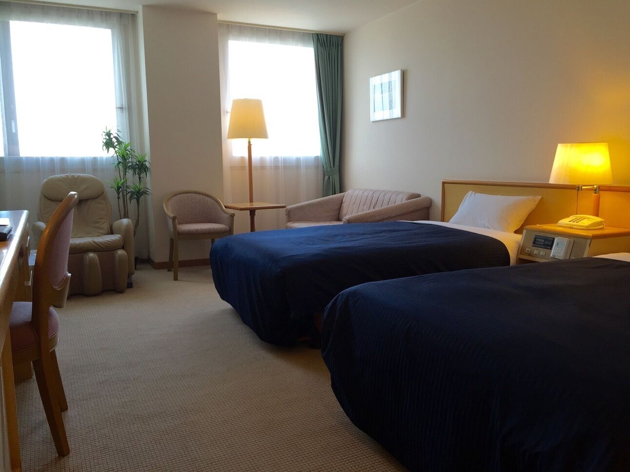 A hotel room with two beds, a desk, a chair, a lamp, and a bedside table.
