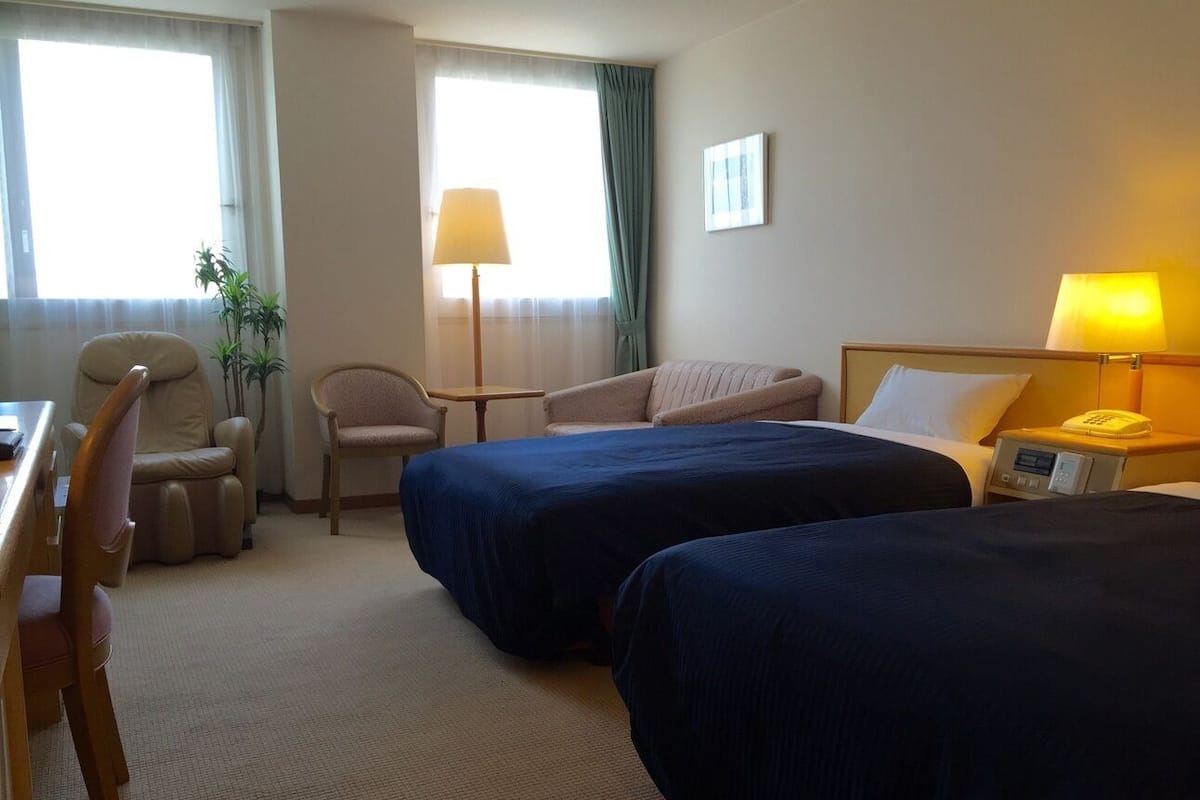 A hotel room with two beds, a desk, a chair, a lamp, and a bedside table.