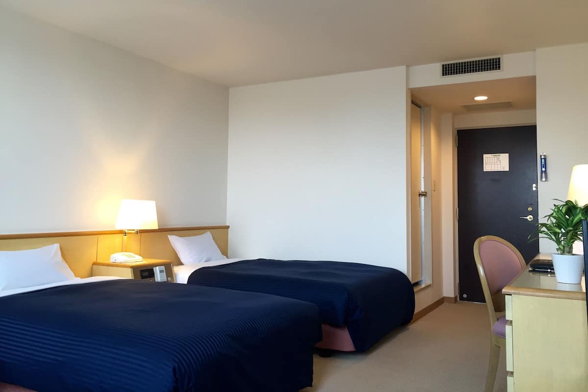 A hotel room with two beds, a desk with a chair, a lamp, and a potted plant.