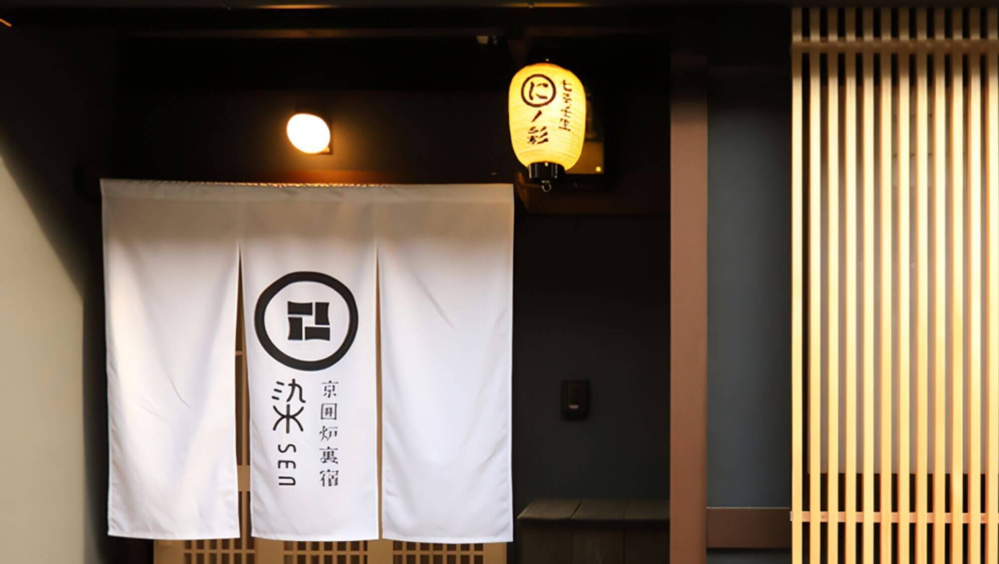 Ein Zimmer mit weißem Vorhang, einem Logo und japanischem Text, einer traditionellen japanischen Laterne und einer Schiebetür.