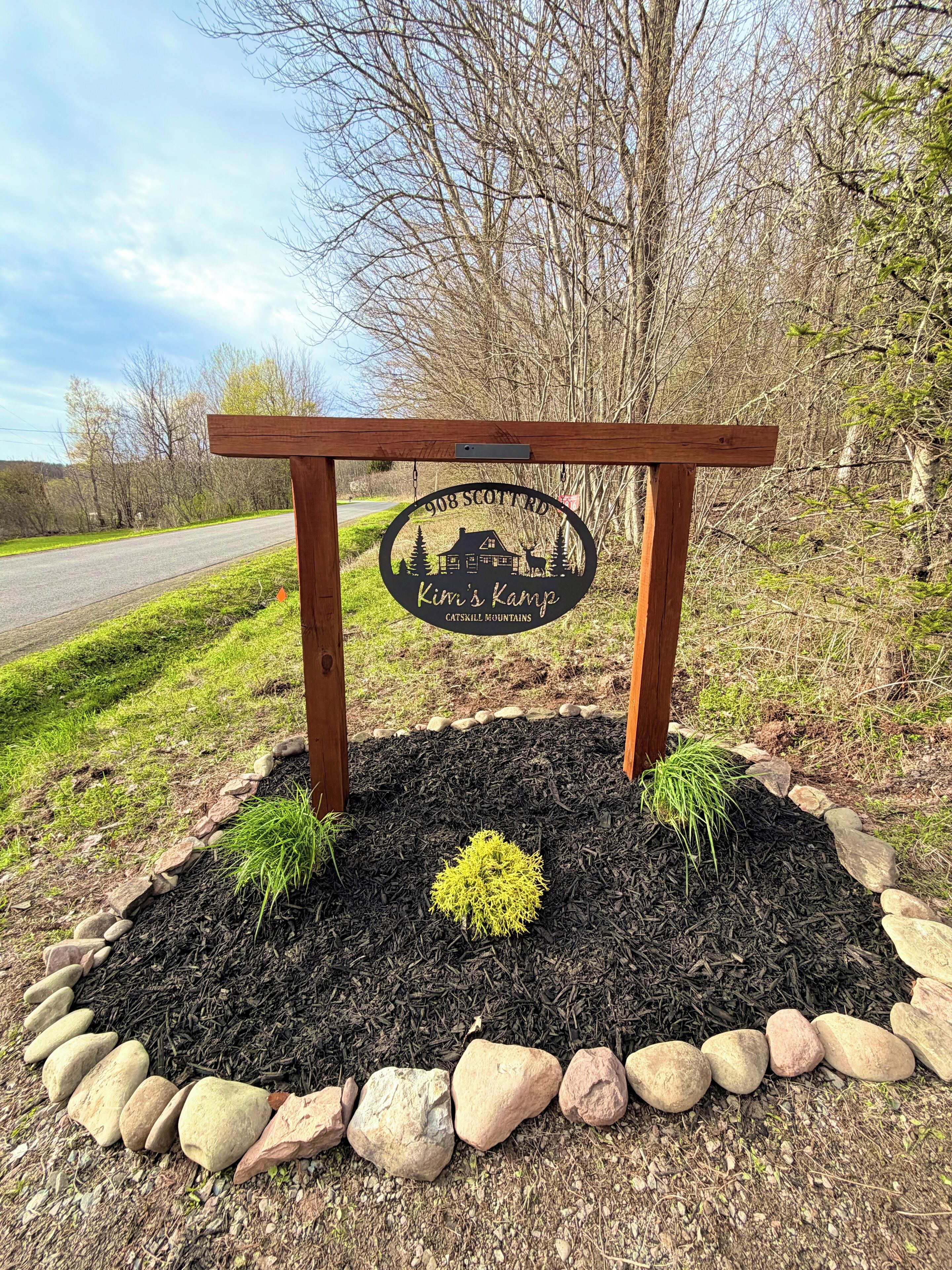 A wooden sign with a house and trees, surrounded by a rock and grass border.