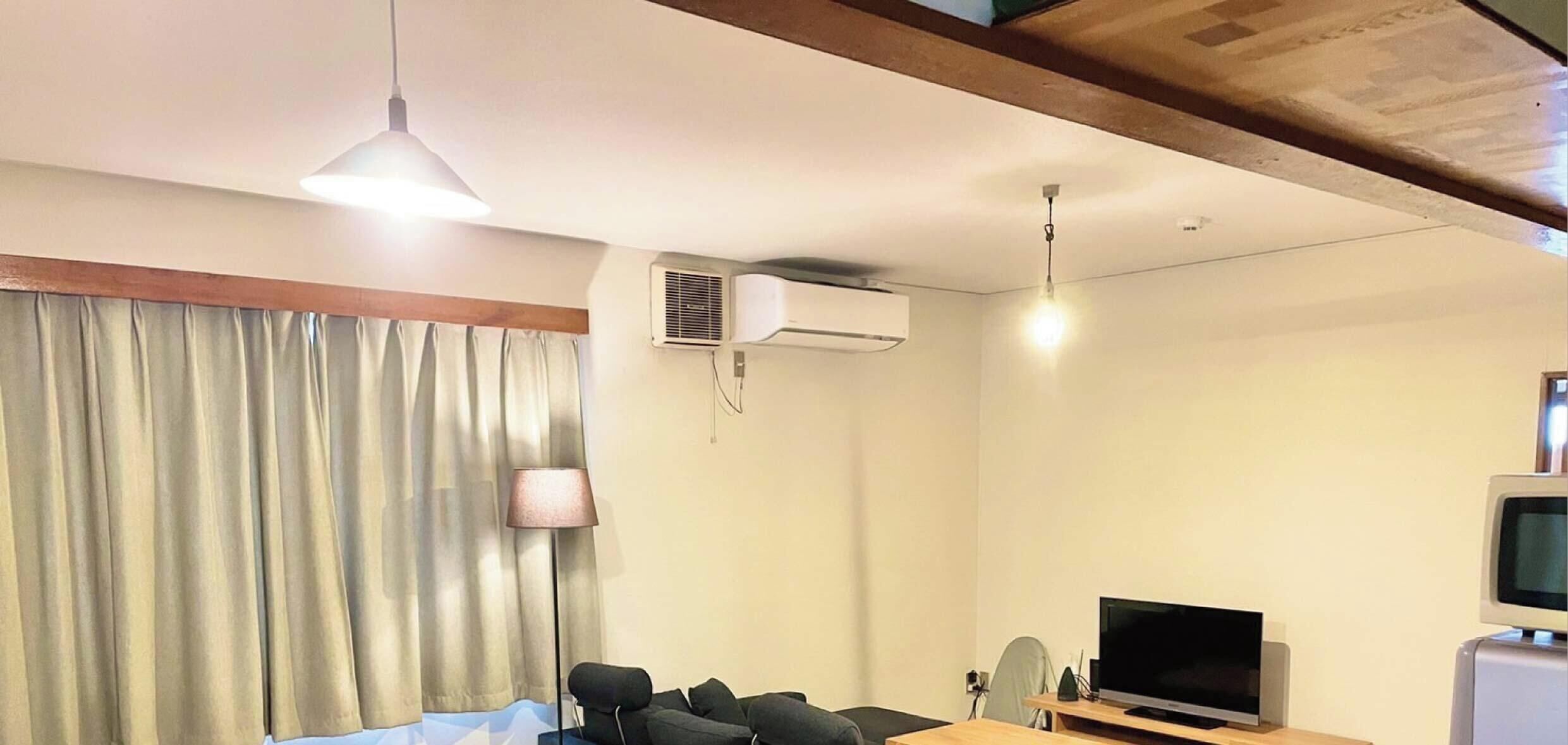 A hotel room with a bed, a desk with a computer, a television, an air conditioning unit, and a window with curtains.