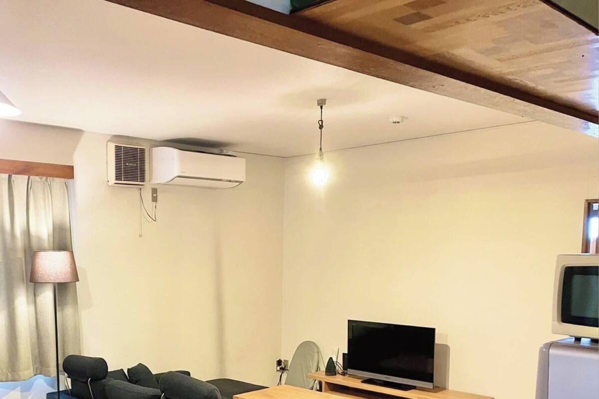 A hotel room with a bed, a desk with a computer, a television, an air conditioning unit, and a window with curtains.