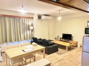 A modern living room with a sofa, a dining table, a TV, and a small kitchen area. - Tsubaki House (Hakodate)