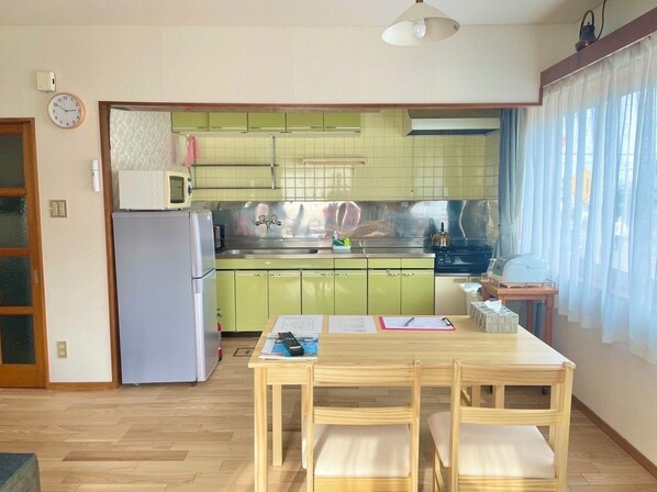 A compact kitchen with a refrigerator, microwave, and stove, a dining table with chairs, and a window with curtains. - Tsubaki House (Hakodate)