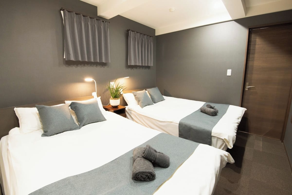 Une chambre d’hôtel avec deux lits, des rideaux gris, une plante en pot et une porte en bois.