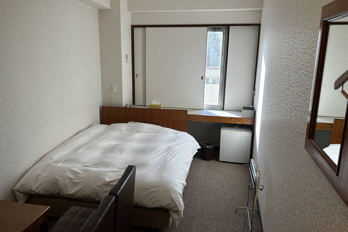 Una habitación de hotel compacta con una cama, un pequeño escritorio, un espejo y una ventana.