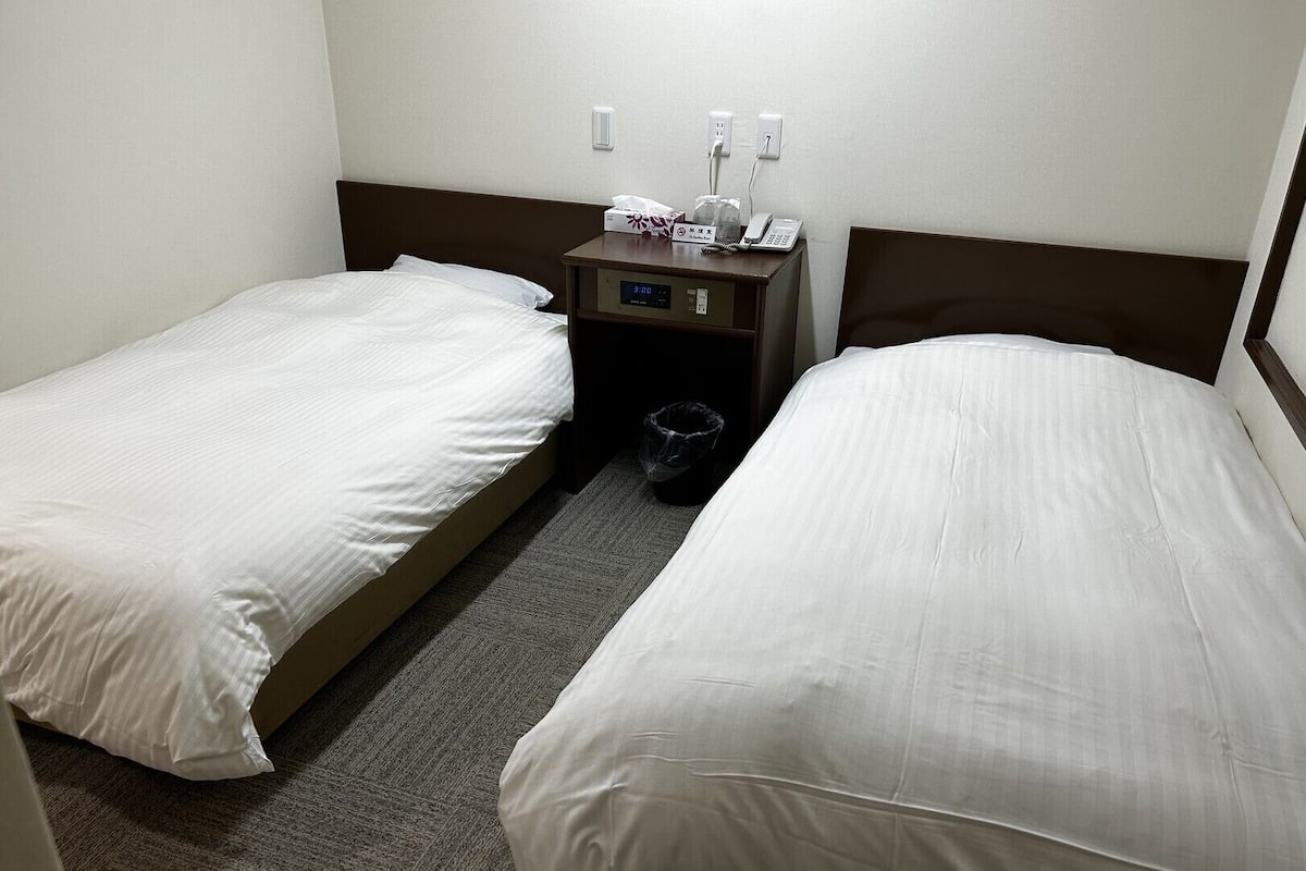 Habitación de hotel con dos camas individuales, una mesita de noche con teléfono y reloj digital, y un cubo de basura.