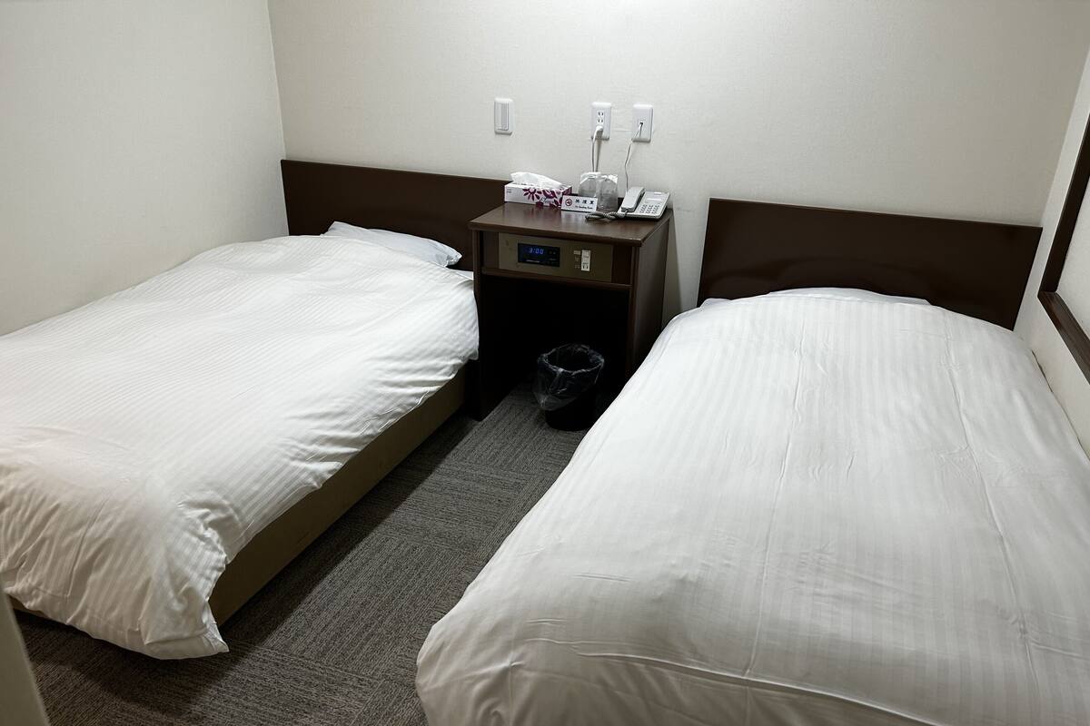 Habitación de hotel con dos camas individuales, una mesita de noche con un teléfono y un reloj digital, y un cesto de basura.