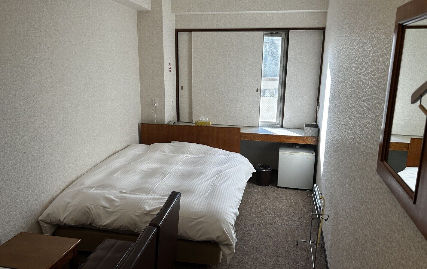 Una habitación de hotel compacta con una cama individual, un escritorio pequeño, un espejo, una ventana y un mini refrigerador.