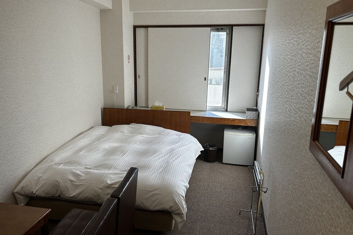 Una habitación de hotel compacta con una cama individual, un escritorio pequeño, un espejo, una ventana y un mini refrigerador.